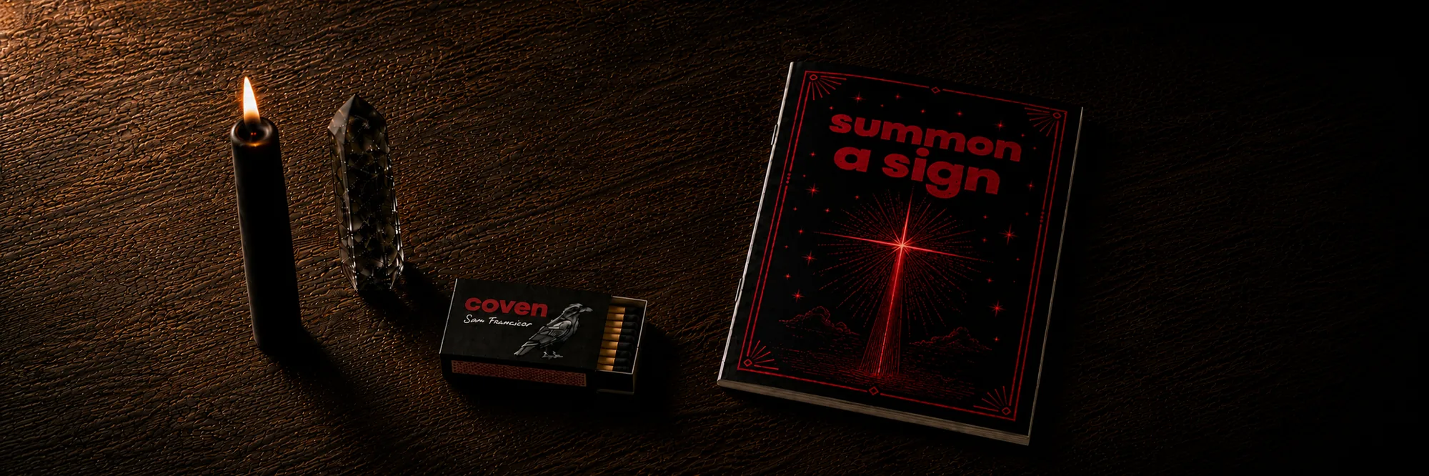 Four Coven objects arranged on a dark wood surface: a lit black candle, a smoky quartz crystal point standing upright, an open Coven-branded matchbox with red-tipped matches, and a Summon a Sign spellbook with red ink illustration of a beam of light