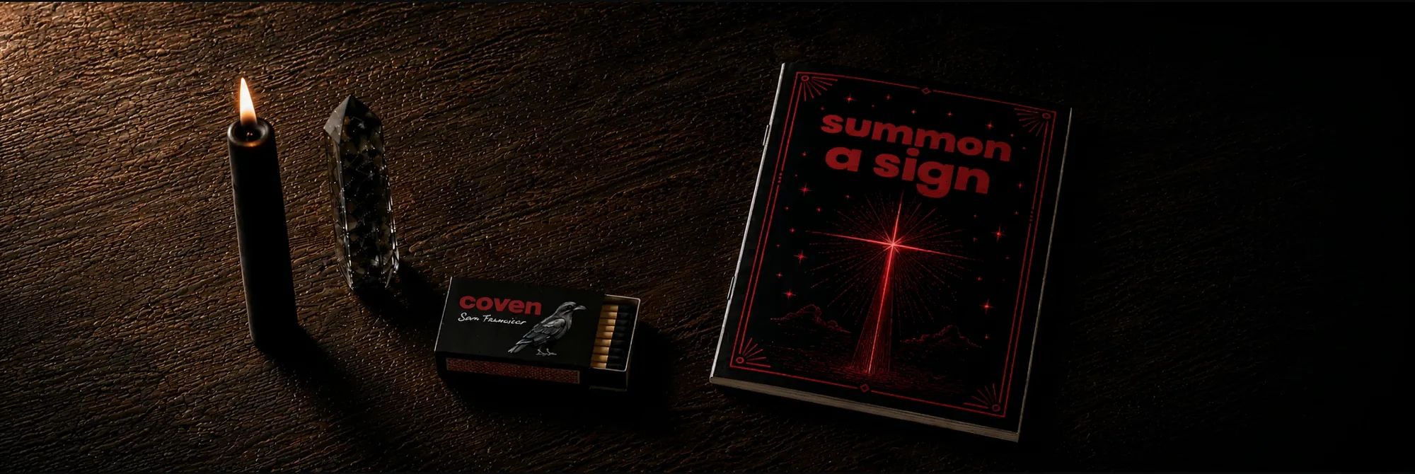 Four Coven objects arranged on a dark wood surface: a lit black candle, a smoky quartz crystal point standing upright, an open Coven-branded matchbox with red-tipped matches, and a Summon a Sign spellbook with red ink illustration of a beam of light
