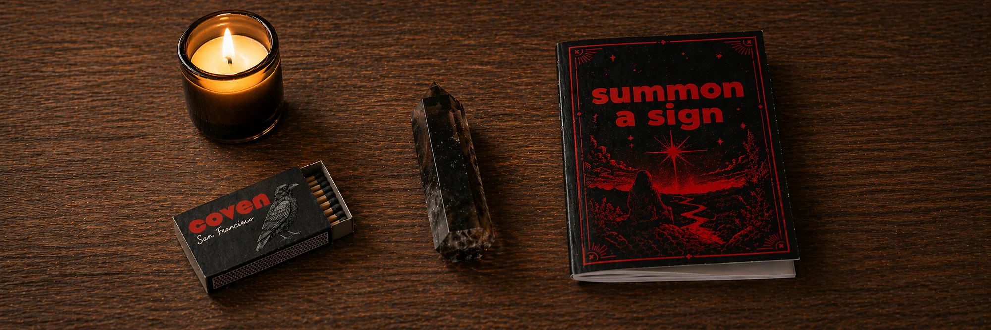 Five Coven objects arranged on a dark wood surface: a Coven-branded matchbox open with matches, a lit candle in a brass-rimmed amber vessel, a smoky quartz crystal point standing upright, and a Summon a Sign spell card with red ink illustration of a figure beneath a star