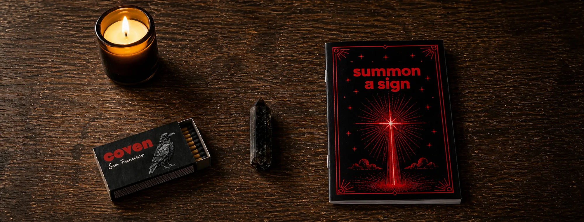 Four Coven objects arranged on a dark wood surface: a lit amber-glass candle, a Coven-branded matchbox open with matches, a smoky quartz crystal point standing upright, and a Summon a Sign spellbook with red ink illustration of a beam of light over water
