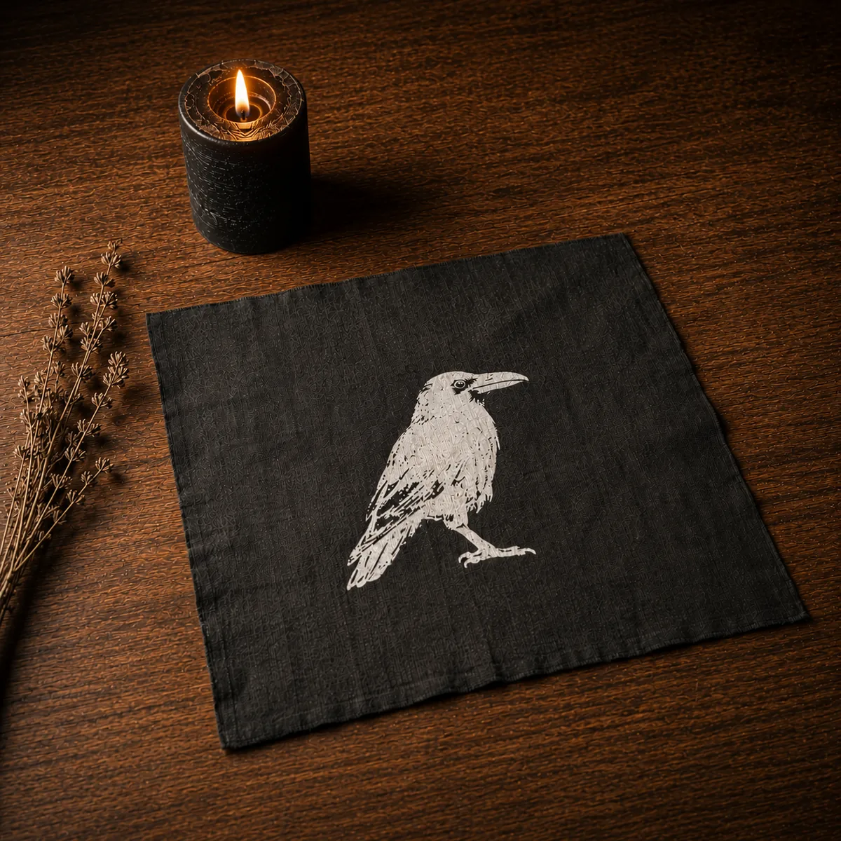 The altar cloth — a heavyweight black linen square hand-screen-printed in bone white with Zelda the raven at center, photographed at an angle on a dark wood surface beside a lit black pillar candle and a sprig of dried herbs