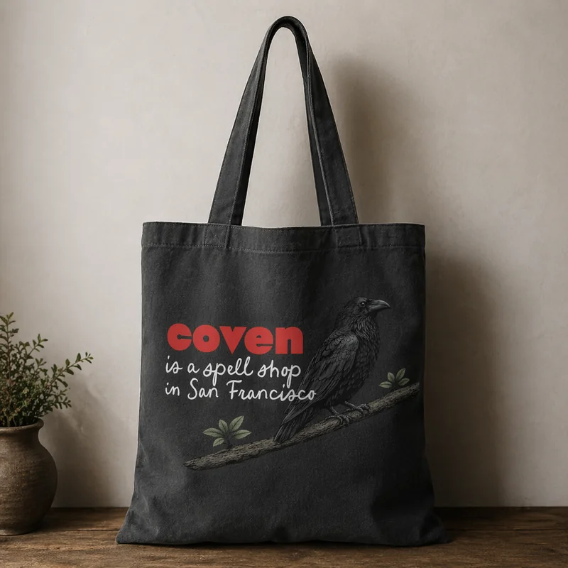 The Carryall — a black canvas tote bag with the Coven wordmark in red and an illustrated raven perched on a branch with foliage, propped against a wall beside a potted plant