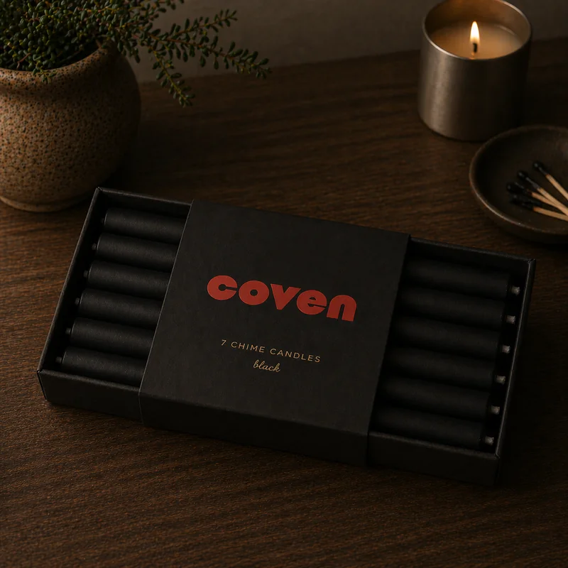 Chime candles — a black box of seven hand-dipped black chime candles with a Coven wordmark in red and '7 chime candles / black' in copper on the sleeve, photographed on a dark wood surface beside a stoneware plant pot, brass candle, and dish of black-tipped matches