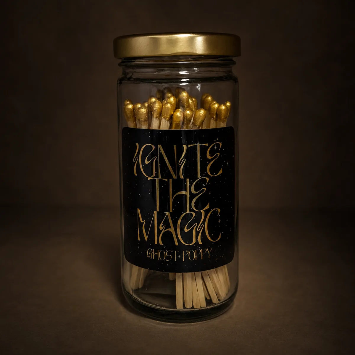 Fancy matches — a glass apothecary jar of gold-tipped matches with a brass lid and a black label reading ‘Ignite the Magic — Ghost Poppy’, on a moody brown backdrop
