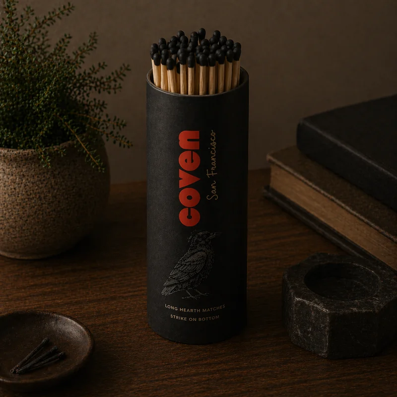 Hearth matches — a tall black cylindrical tube of long matches with the Coven wordmark in red, 'San Francisco' in cursive, an illustrated raven, and 'Long hearth matches, strike on bottom' on the front, photographed on a dark wood surface beside a stoneware plant pot, books, and a stone match holder