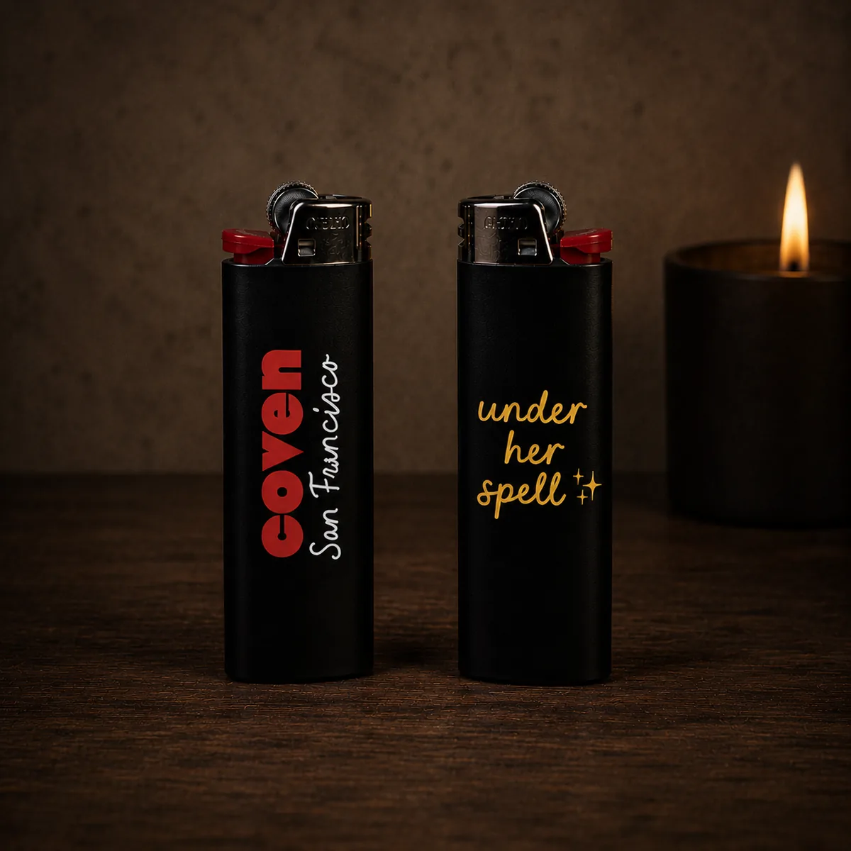 The lighter — a matte black Coven lighter shown front and back. One side reads 'coven' in red with 'San Francisco' in white cursive; the other side reads 'under her spell' in gold script with sparkles, photographed on a dark wood surface beside a lit black pillar candle in moody candlelight