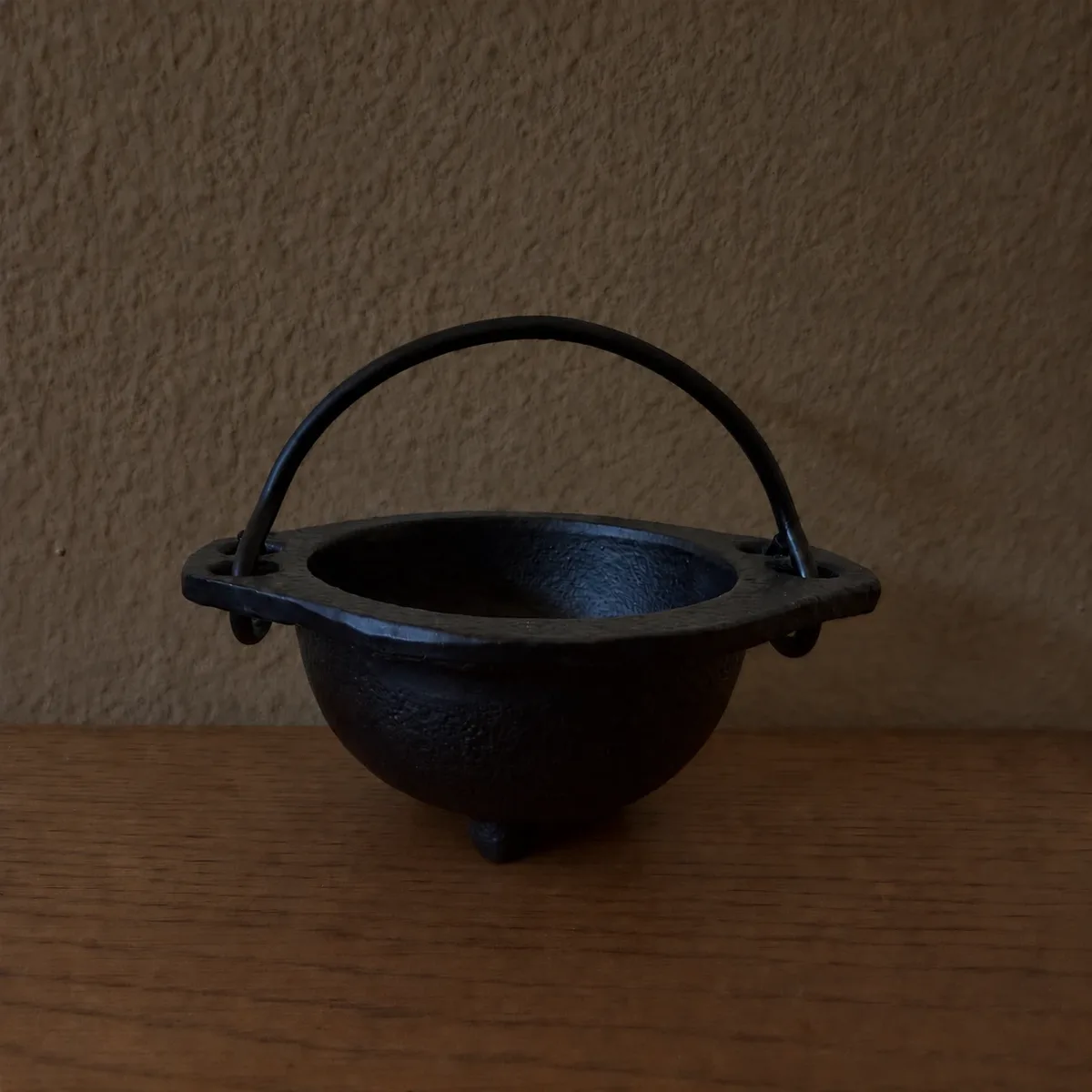 Mini cauldron — a small black cast-iron cauldron with three feet and a wire handle, on a wooden surface against a tan wall