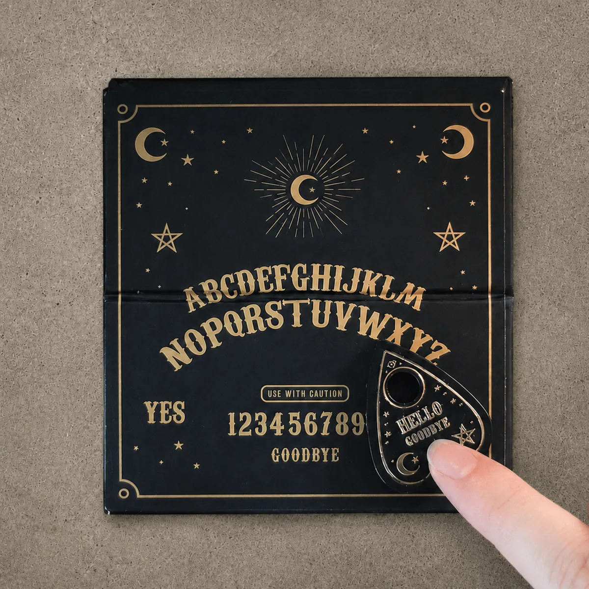 Mini Ouija, second view — the black travel talking board laid open and flat, a finger guiding the gold-and-black planchette across the gold lettering