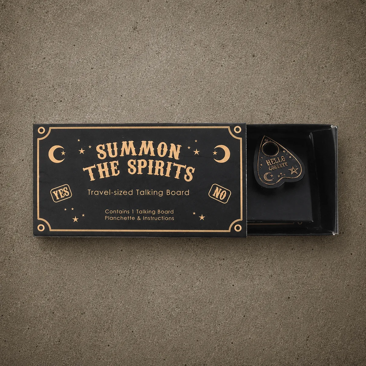 Mini Ouija — a black travel-sized talking board with gold lettering — the alphabet, numbers, sun and moon symbols, and yes/no — photographed top-down on a warm cork surface with a small gold-and-black planchette