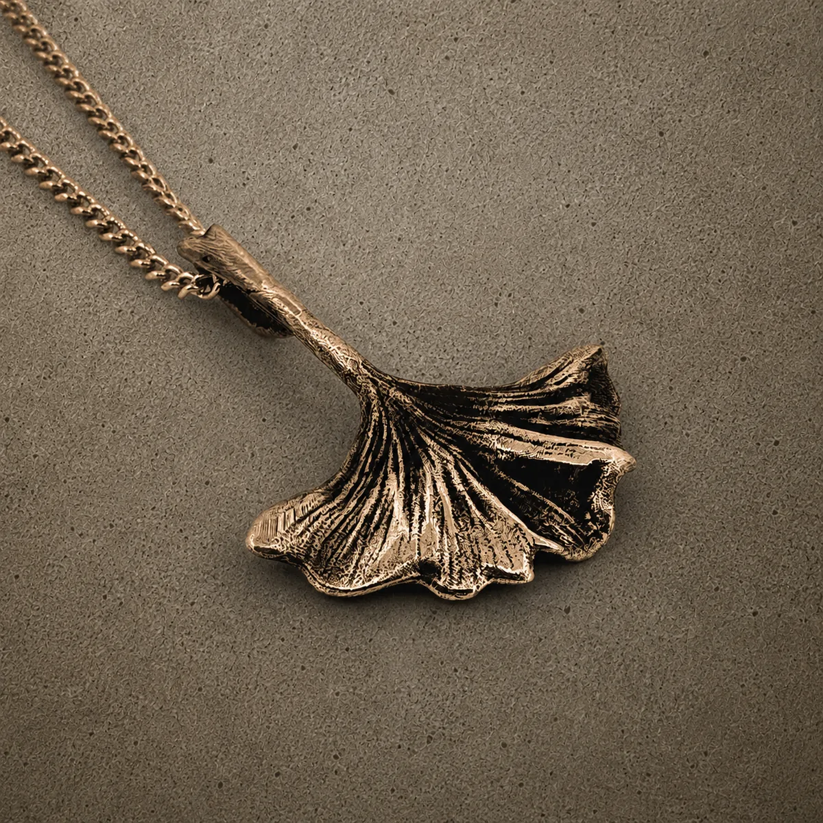 Yellow bronze ginkgo leaf pendant on a chain, photographed against a concrete surface