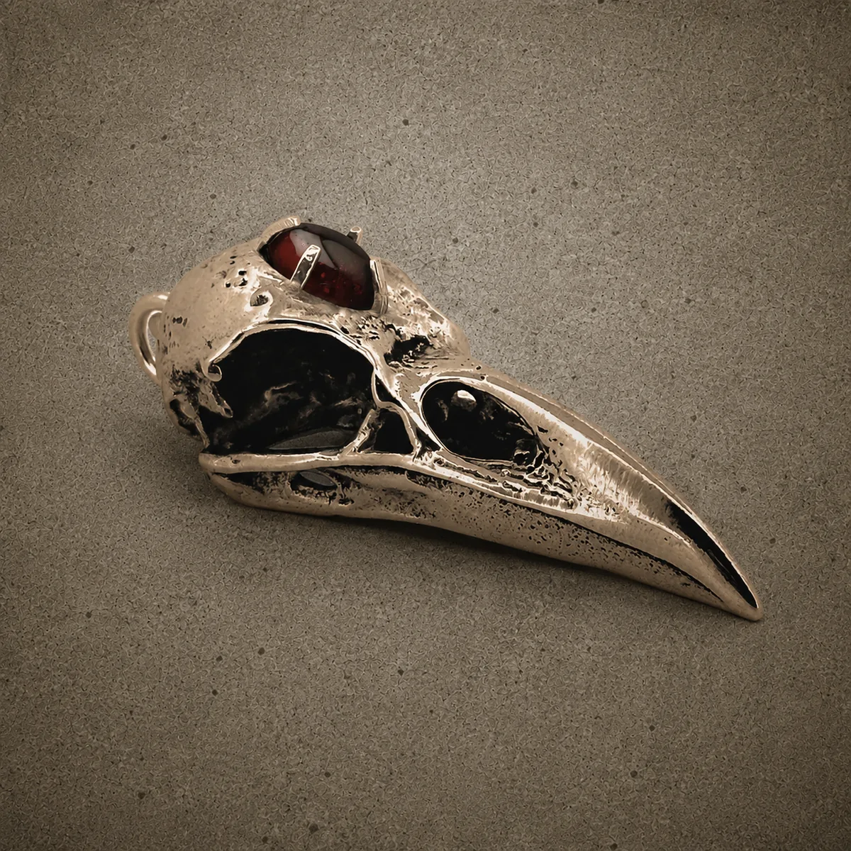 Yellow bronze raven skull pendant inset with a deep-red garnet, photographed against a concrete surface