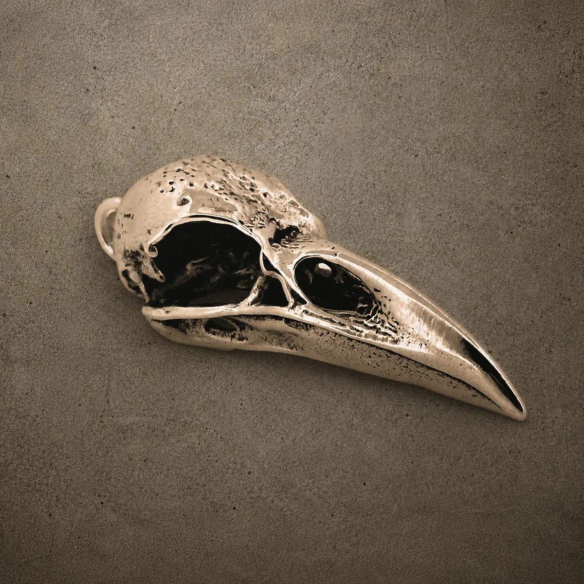 Yellow bronze raven skull pendant with a long curved beak, photographed against a concrete surface