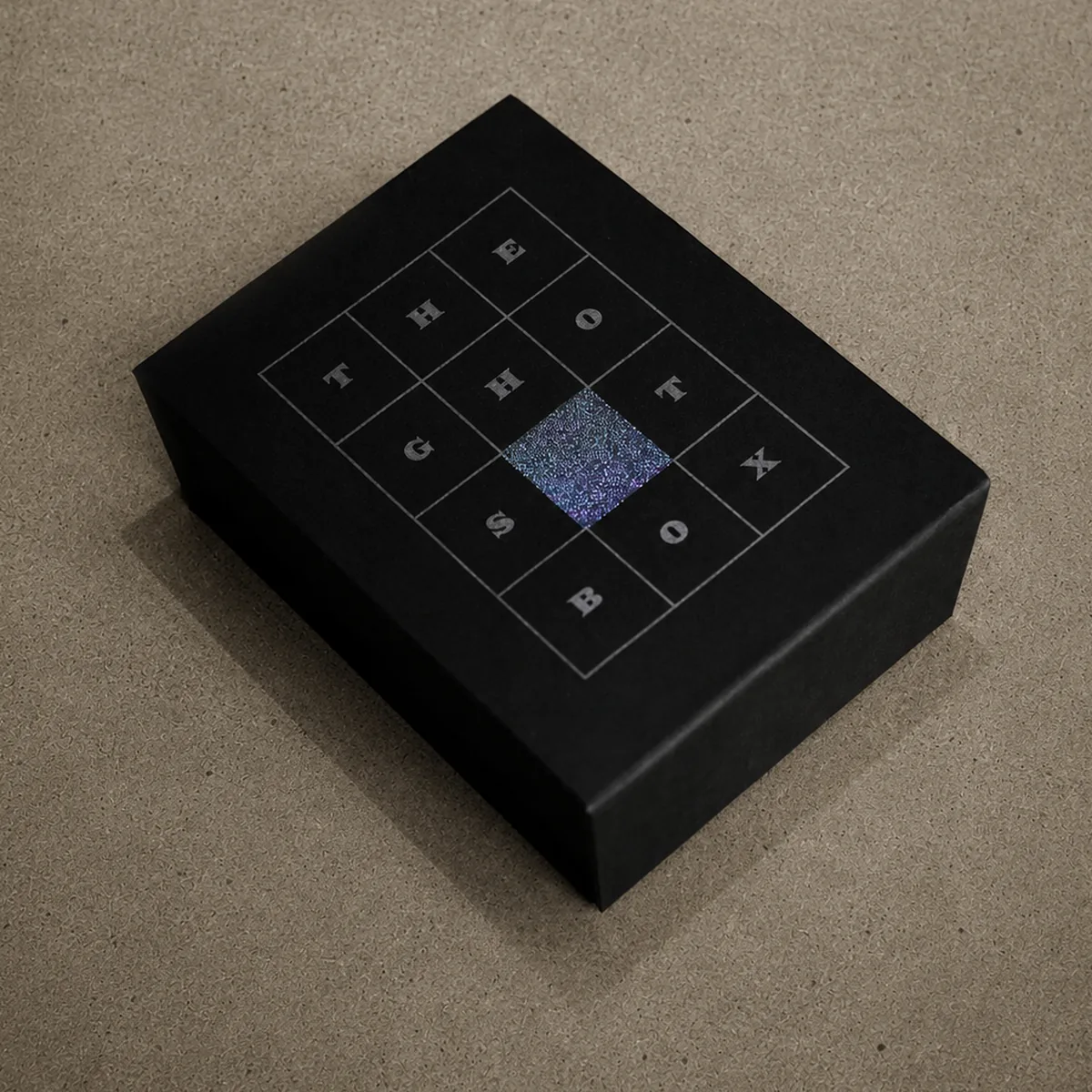 The Ghost Box — a matte black box stamped THE GHOST BOX in a 4x4 grid with a single iridescent tile at its center, on a cork surface