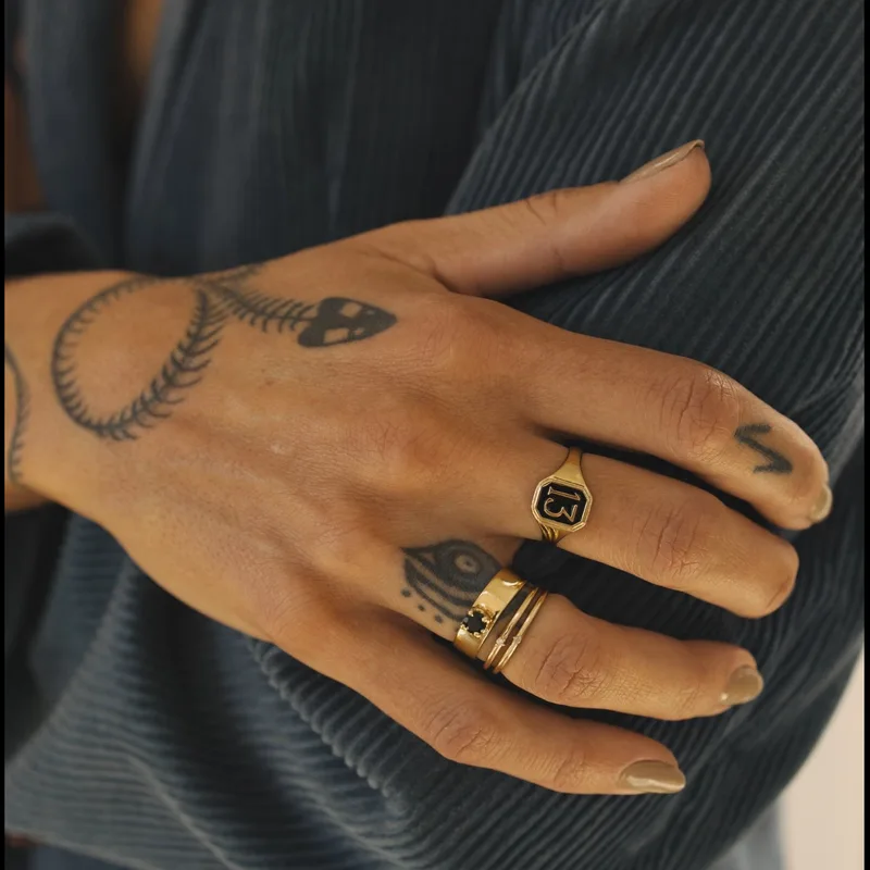 Signet ring — worn on a tattooed hand resting against a ribbed charcoal sweater, stacked with other gold rings