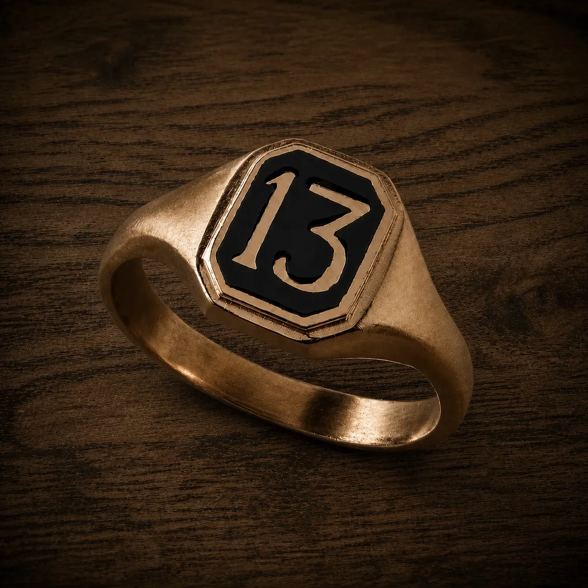 Signet ring — close angled product shot of the gold-plated signet ring with black enamel '13' face, photographed on a warm concrete surface