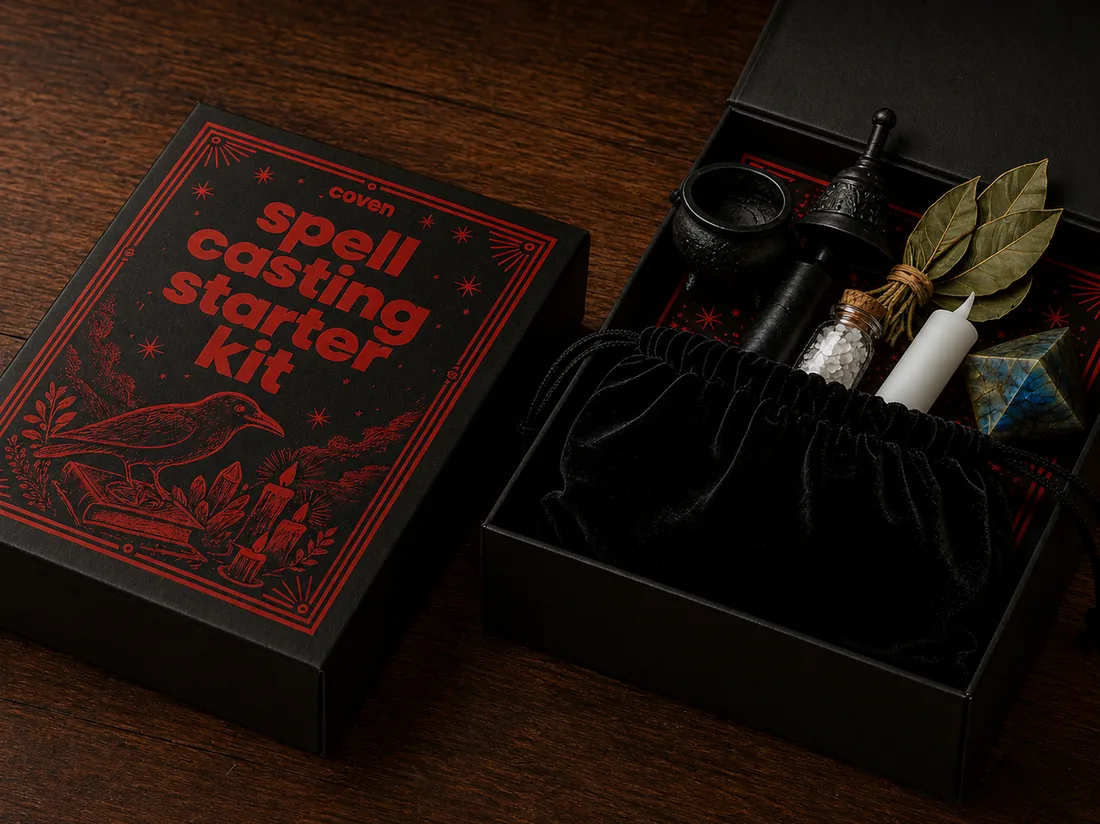 The Spellcasting Starter Kit open at an angle on a dark wood surface — black magnetic box with a red-inked raven illustration, beside a velvet pouch holding salt, a candle, bay leaves, a labradorite pyramid, and a small mortar